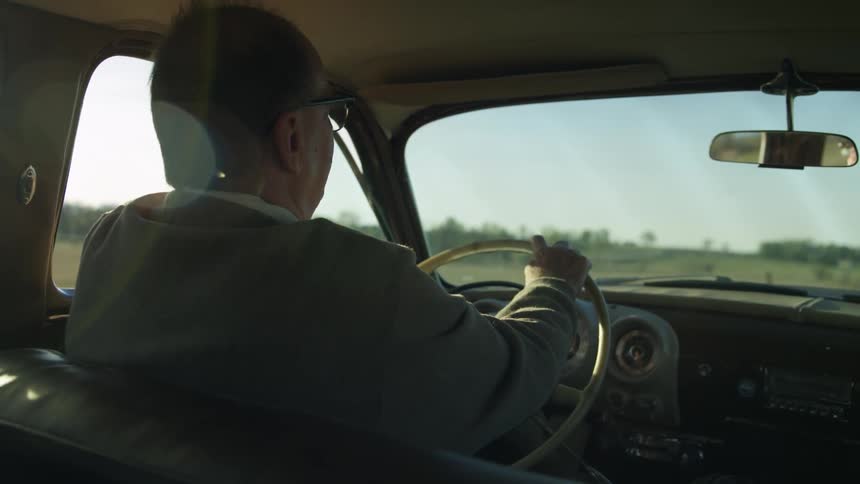 A man drives a vintage car past rural fields on a bright sunny day