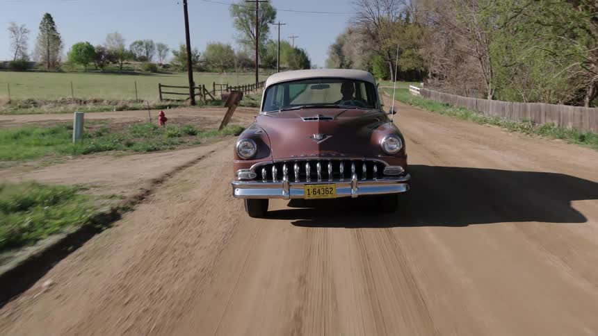 Vintage car drives along dirt road past pastures and mailboxes