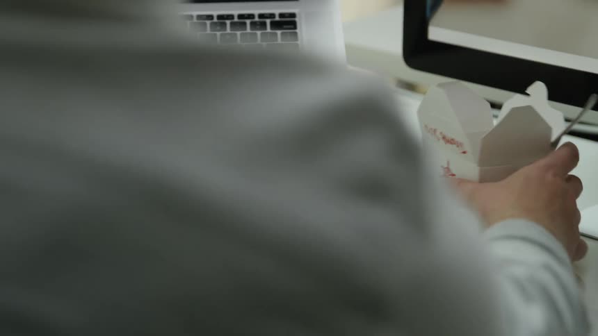 A man sits and types on a keyboard at his desk and then picks up a container of food