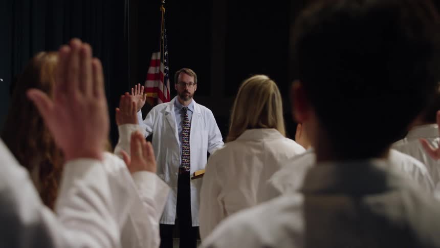 A man stands in front of a group of students all wearing white coats with one hand up taking an oath