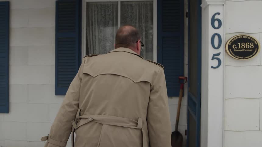 A man in a trench coat walks toward a house