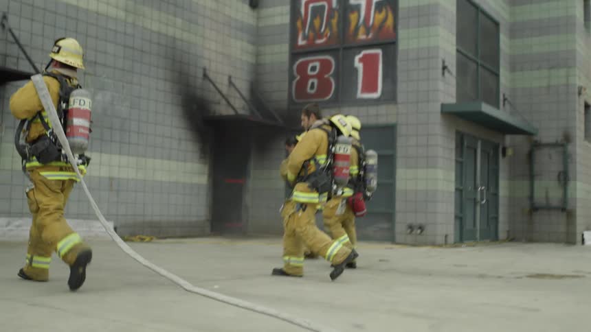 Firefighters in a training exercise runs towards a building carrying hoses and equipment