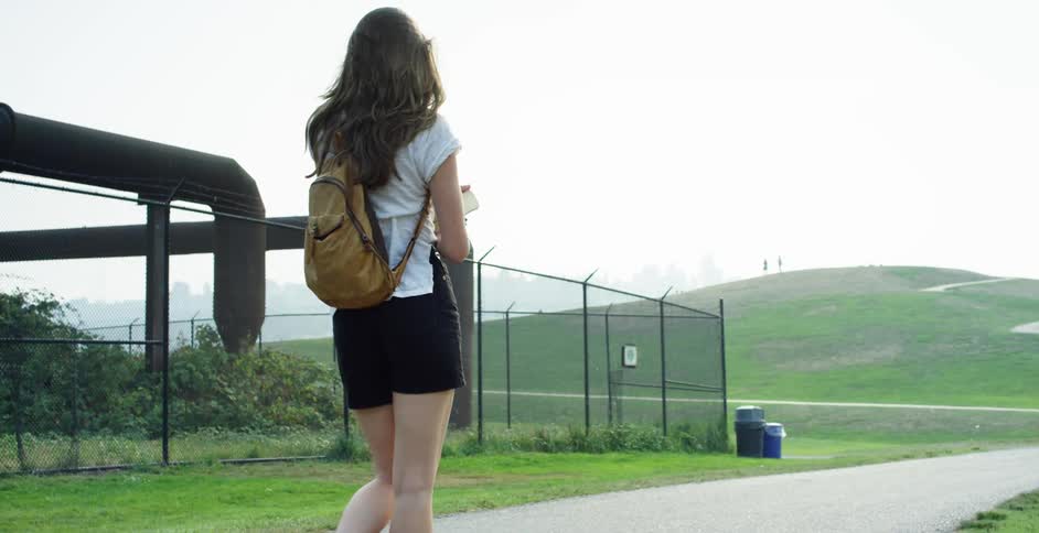 Woman with black shorts and brown backpack holds a book and walks along a narrow road