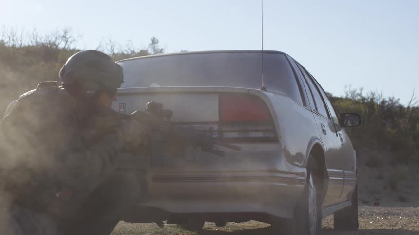 A police officer in tactical gear fires a rifle from behind a car