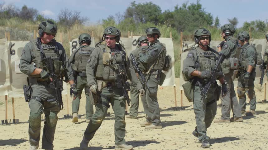 Police officers with helmets shades and rifles walking with targets and trees behind them