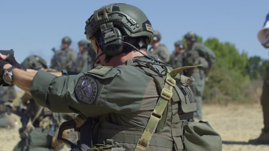A police officer in tactical gear stands outside with other cops and fires a pistol