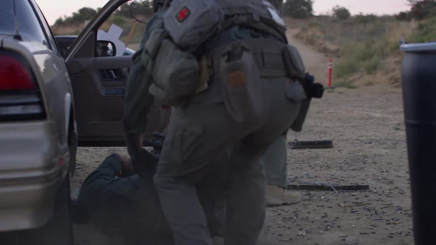 A medic runs to take care of a wounded soldier by a car as another military man provides cover fire