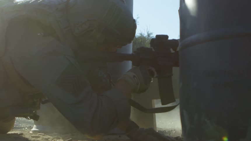 A soldier in full military gear shoots a rifle from behind two barrels