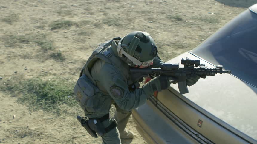 Police officer in military gear takes cover behind a car and takes aim with an assault rifle