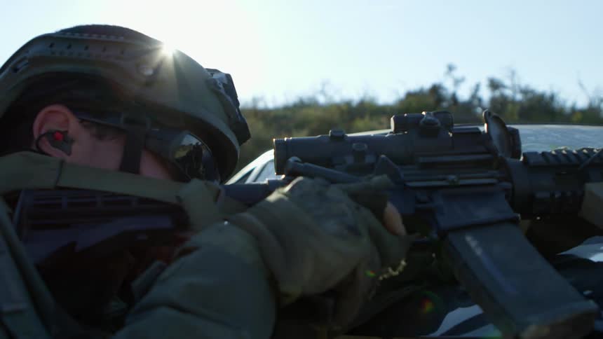 A police officer in a tactical gear leans over a car and fires a rifle