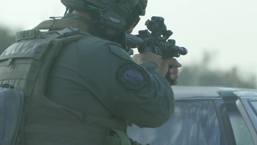 A police officer in gear shoots a rifle over the top of a parked car