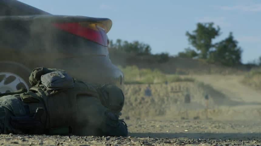 A police officer lies on the ground and takes cover behind a vehicle while shooting at a target