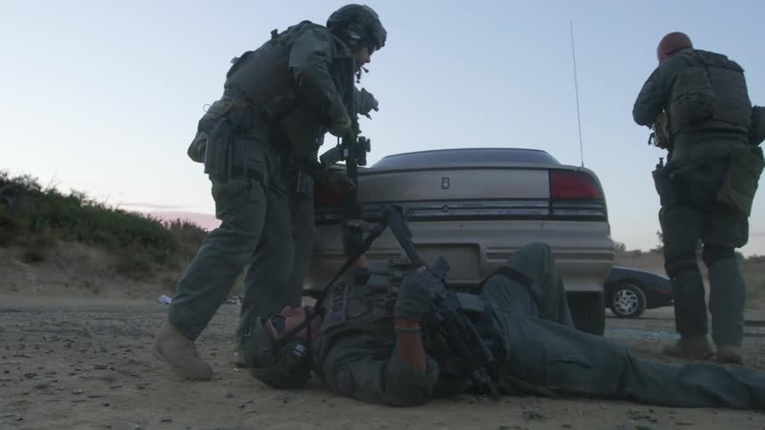 A swat team member pulls another officer to safety behind a car