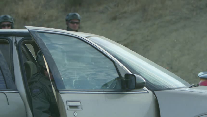 Soldiers stand around a car as shots are fired and another soldier gets out of the car with his rifle
