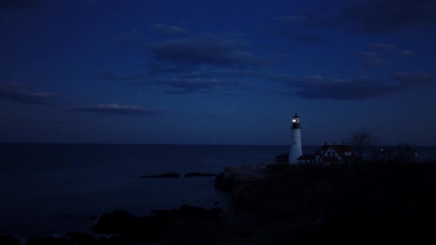 Lighthouse light flashes on promontory overlooking ocean at night under a cloudy sky