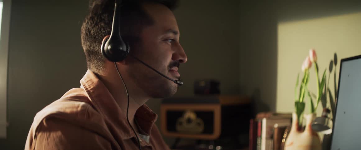 A man wearing a headset talks while drinking coffee and looking at his computer