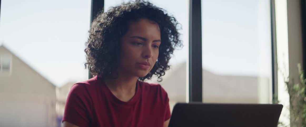 A woman sits at home in front of a large window and uses her laptop