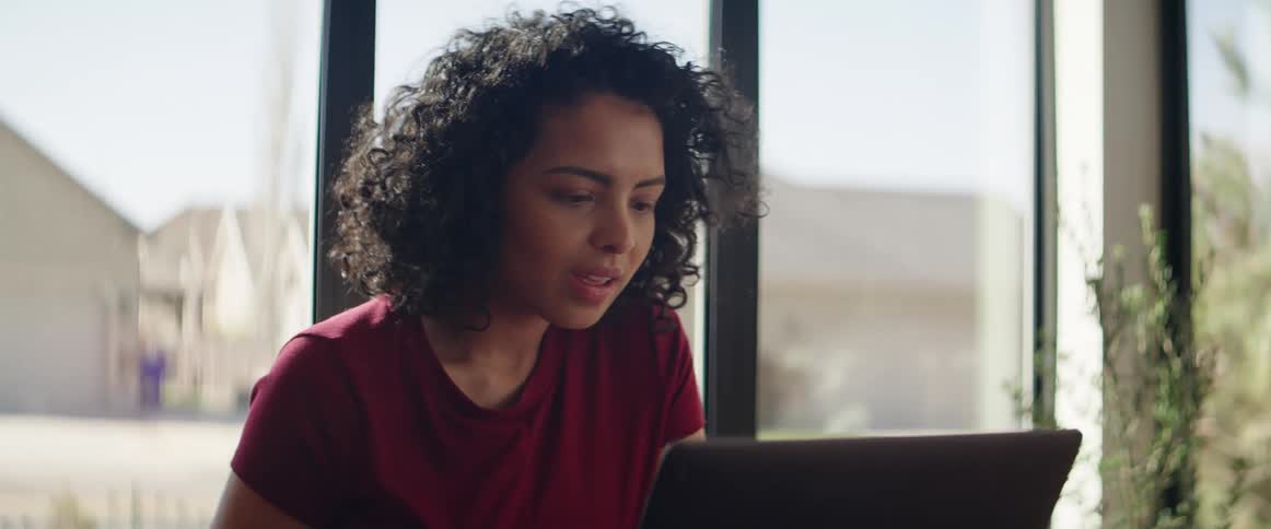 A woman sits at a table in a windowed room while working on her laptop