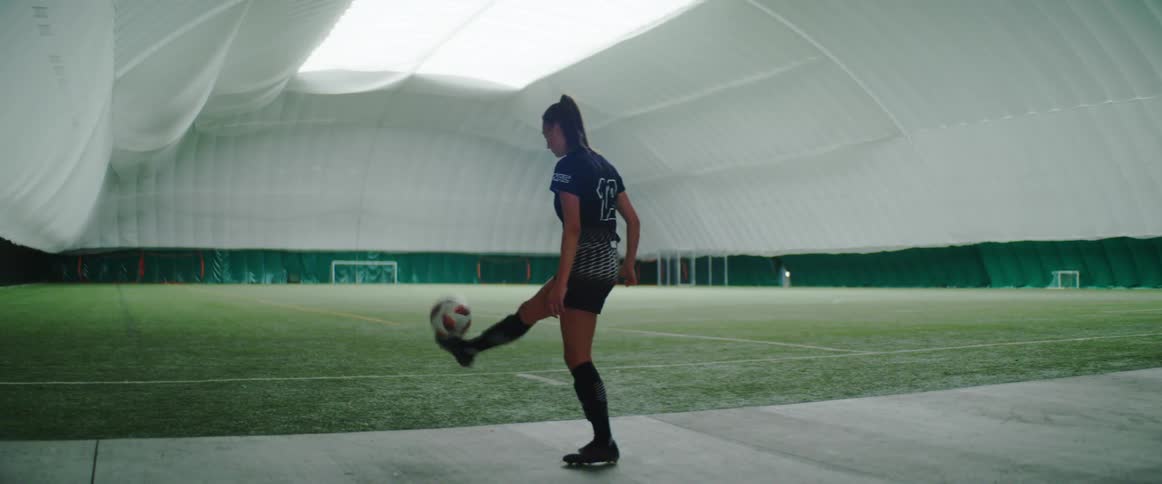A female soccer player is practicing kicking drills while standing on the sidelines of an indoor soccer field