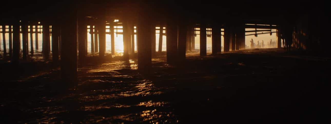 Waves crash on beach under silhouetted pier at sunset