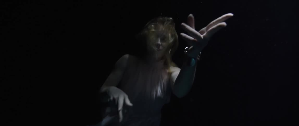A blonde white woman in a dress swims underwater while gesturing with her hands