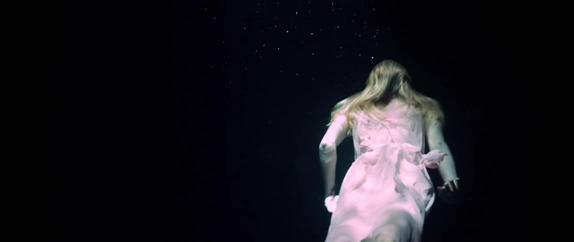 A woman with blonde hair and wearing a sheer gown floats and then swims under black water with a light shining on her