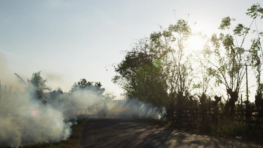 A man drives a motorcycle down a dirt road with a fire burning