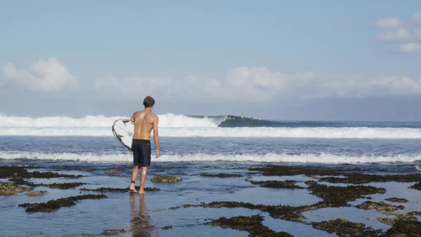 A white surfer with his board stands on a beach and watches the ocean