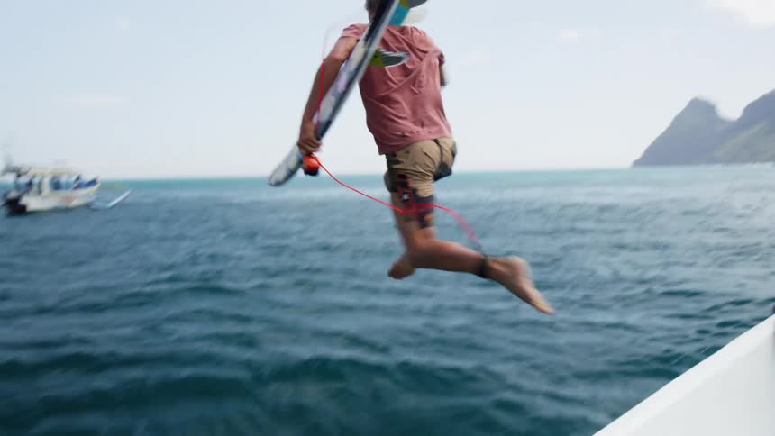 Man jumps off boat into ocean with surfboard as another boat floats nearby