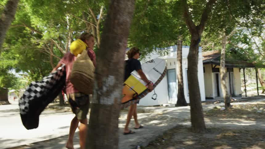Two teenagers carrying their surfboards along a sidewalk as children play