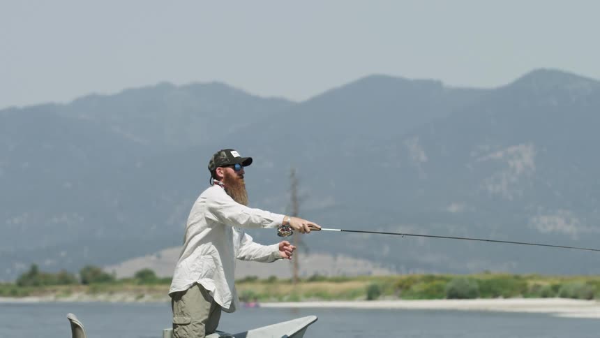 A white man stands in a boat and casts a fishing line into a lake