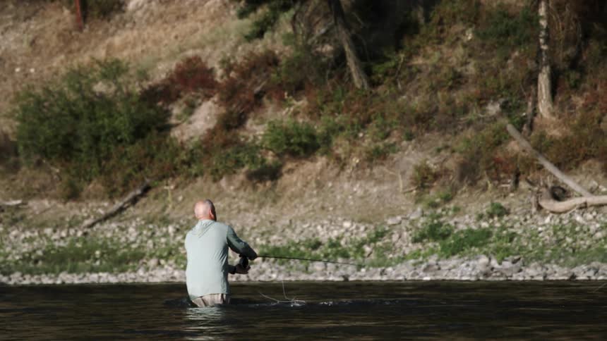 Man stands in river casting fishing line toward steep slope of shore