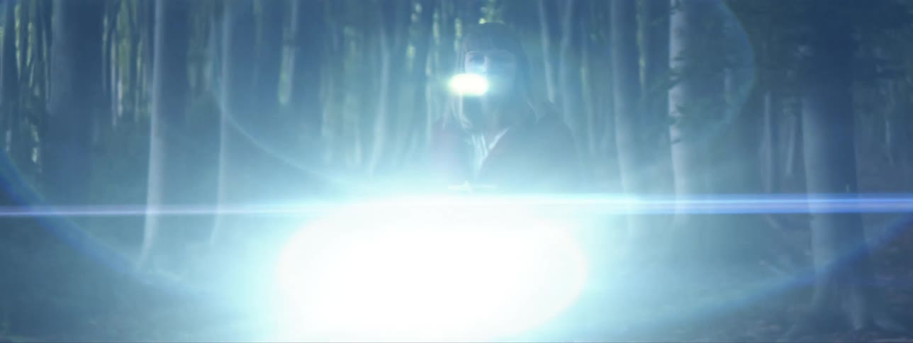 A young woman stands in a dark forest and shines a flashlight as she looks around