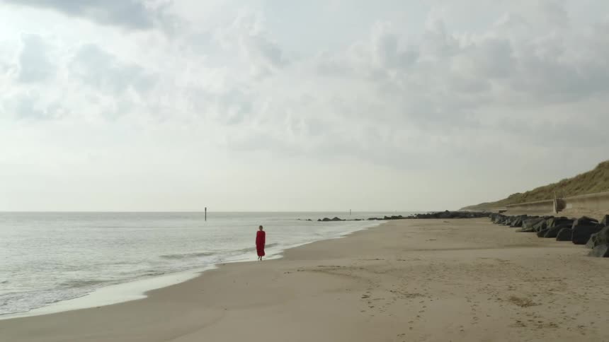 A woman in a red dress walks along a beach just out of reach of the waves