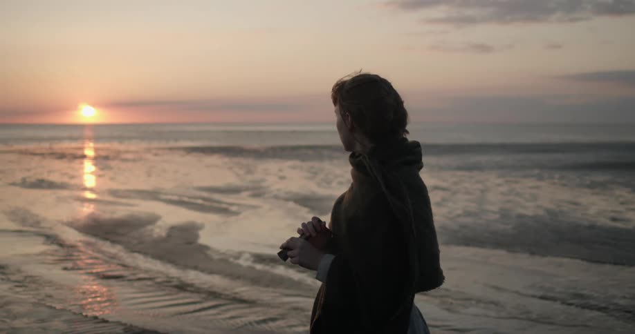 Woman standing on the wet sands of a beach looks at sunset over the ocean