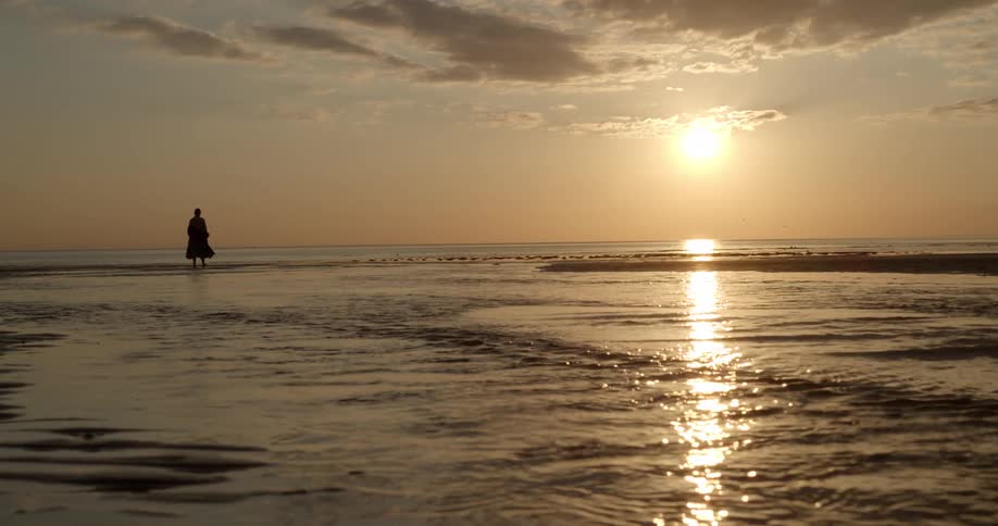 A woman in a long dress wades into the ocean at sunset