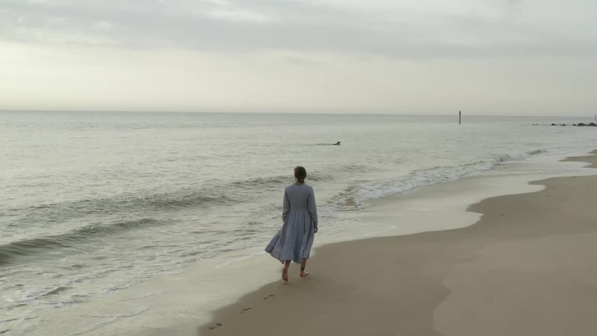 Woman in a dress walks barefoot along the shoreline of the ocean