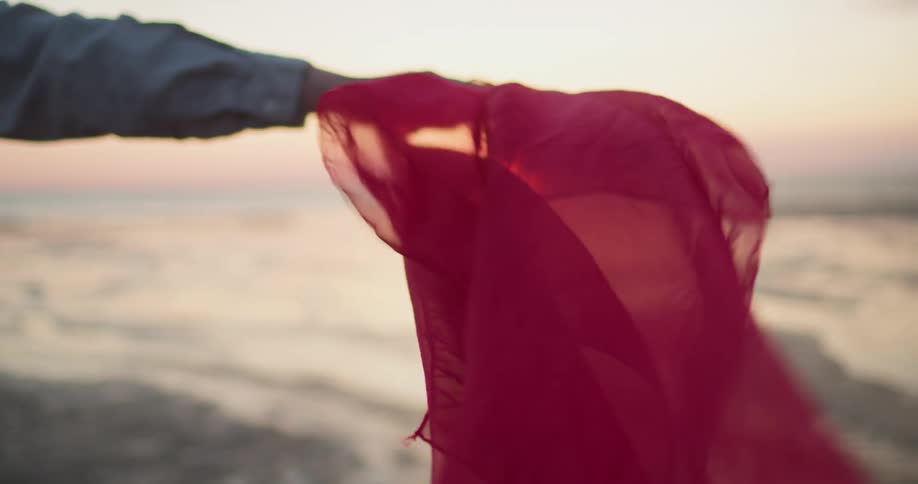 Sun rises on horizon over ocean as person holds fluttering sheer fabric on beach