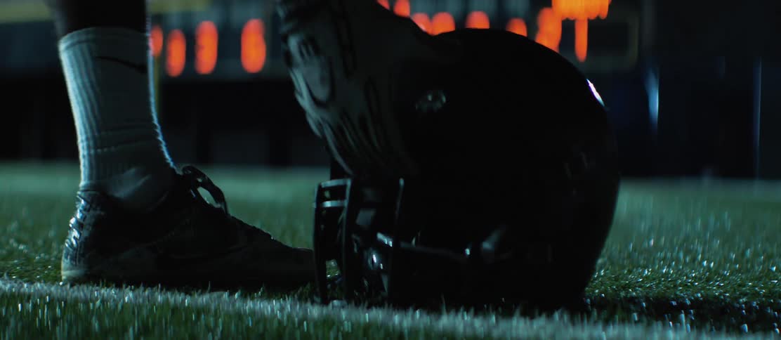 Football helmet lies on a football field at night before a player walks into frame and picks it up