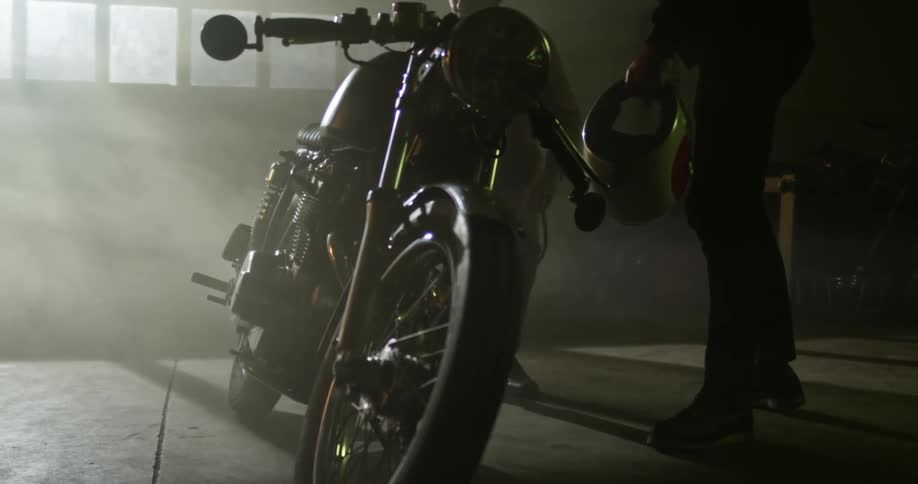 A pair of men examine and walk around a motorcycle in a garage illuminated only by the sunlight coming through the windows