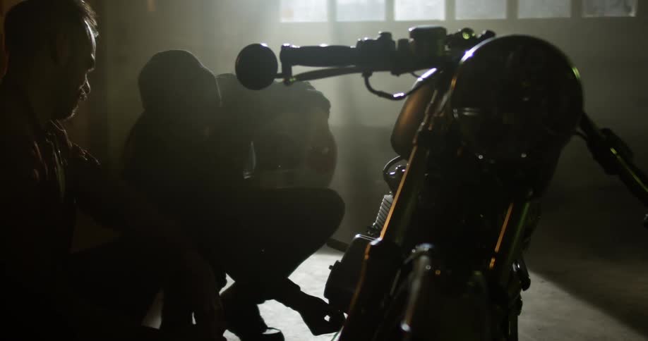 Two men squat in a garage as they check out a motorcycle