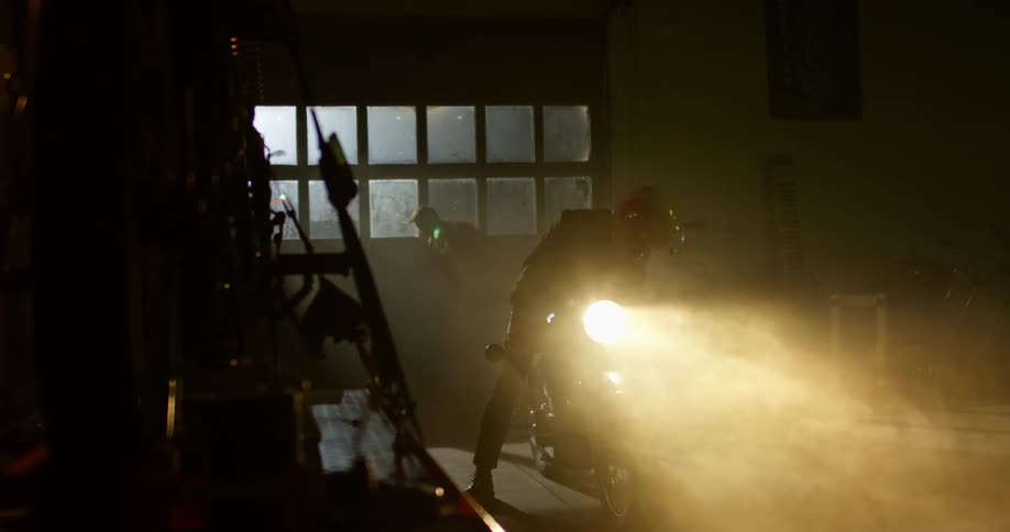 A man starts a motorcycle indoors while his friend closes a garage door