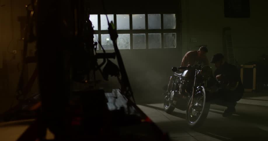Two men working on a motorcycle inside a dimly lit workshop