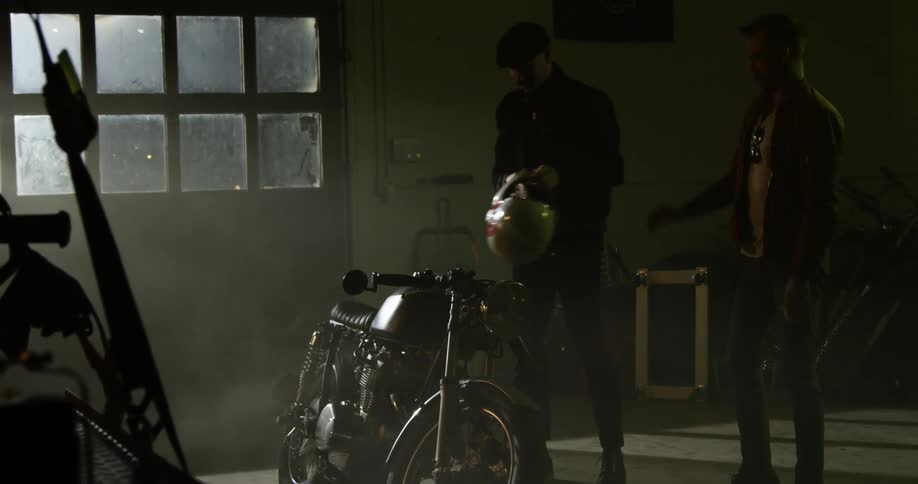 A man handing a helmet to another man as the two approach a motorcycle in a garage