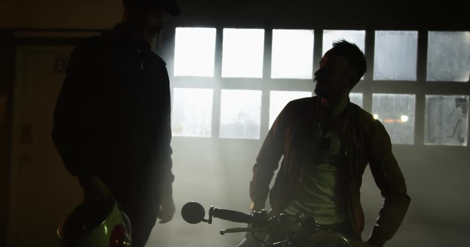 Two man talk to each other around a motorcycle in a dimly lit workshop