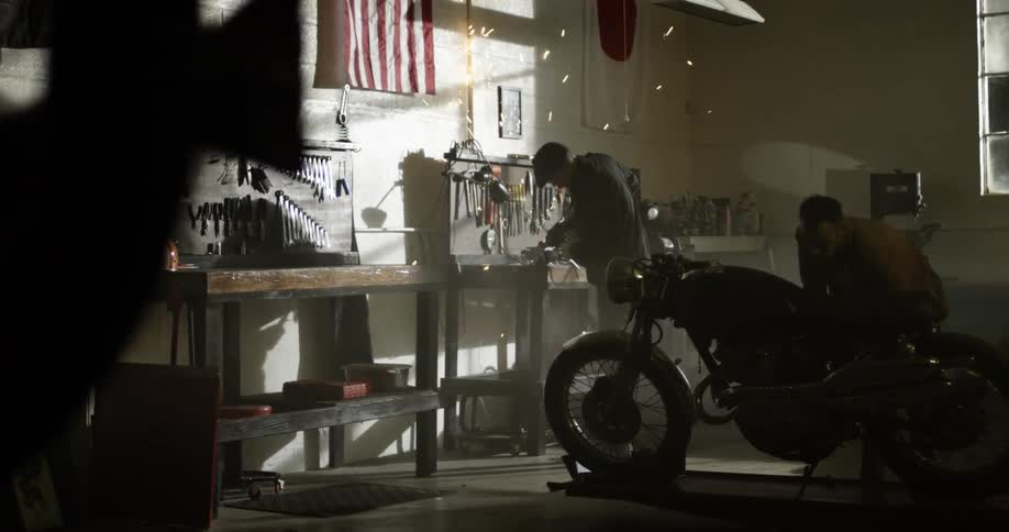 A white man works at a workbench while his friend sits near a motorcycle watching him