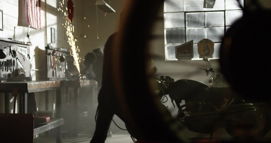 A man welds in a shop as another man walks towards a motorcycle