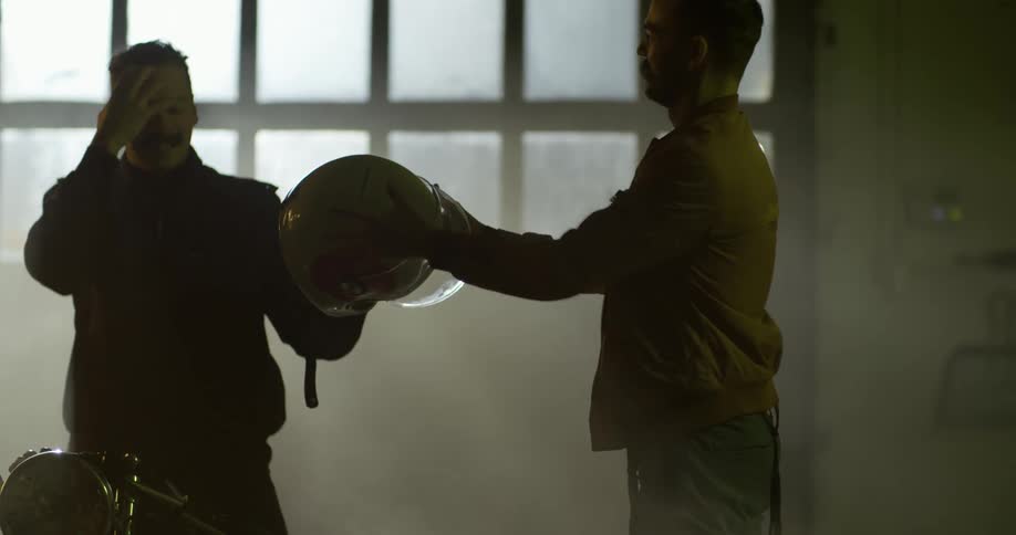 A motorcycle rider takes off his helmet and hands it to his friend before running his hand through his hair