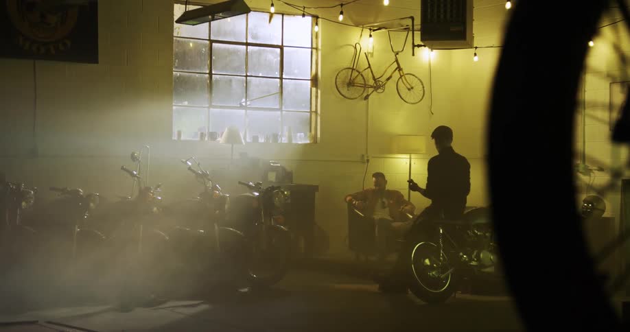 Two men sit in a garage with several motorcycles smoking cigars