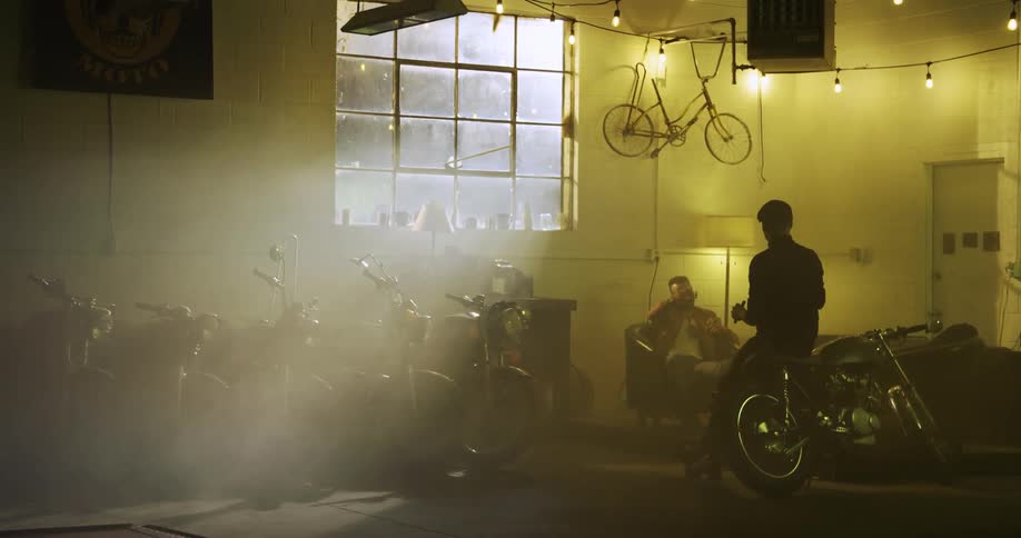 Two men sit and talk in a room filled with motorcycles